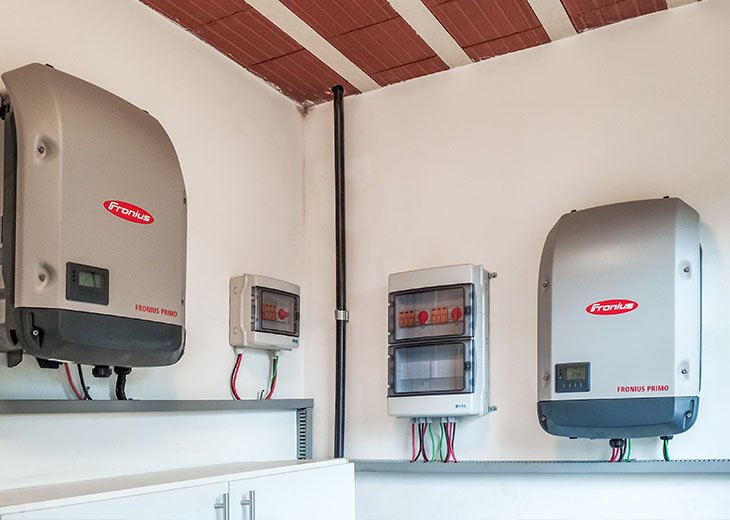 Inversores Fronius instalados em uma parede de uma residência, utilizados para transformar energia solar em eletricidade, com monitores de consumo.