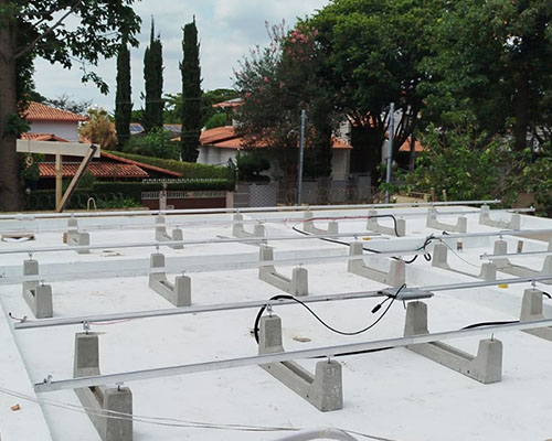 Imagem de estrados metálicos de suporte para instalação de painéis solares em telhado residencial, com árvores e casas ao fundo.