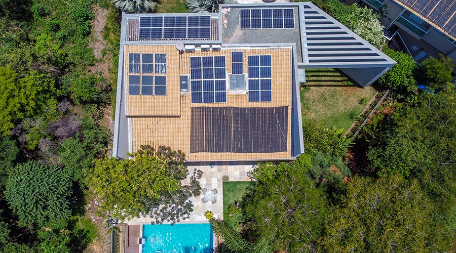 Imagem aérea de uma residência com painéis solares no telhado, cercada por árvores, com piscina ao fundo, mostrando uma instalação de energia solar residencial.